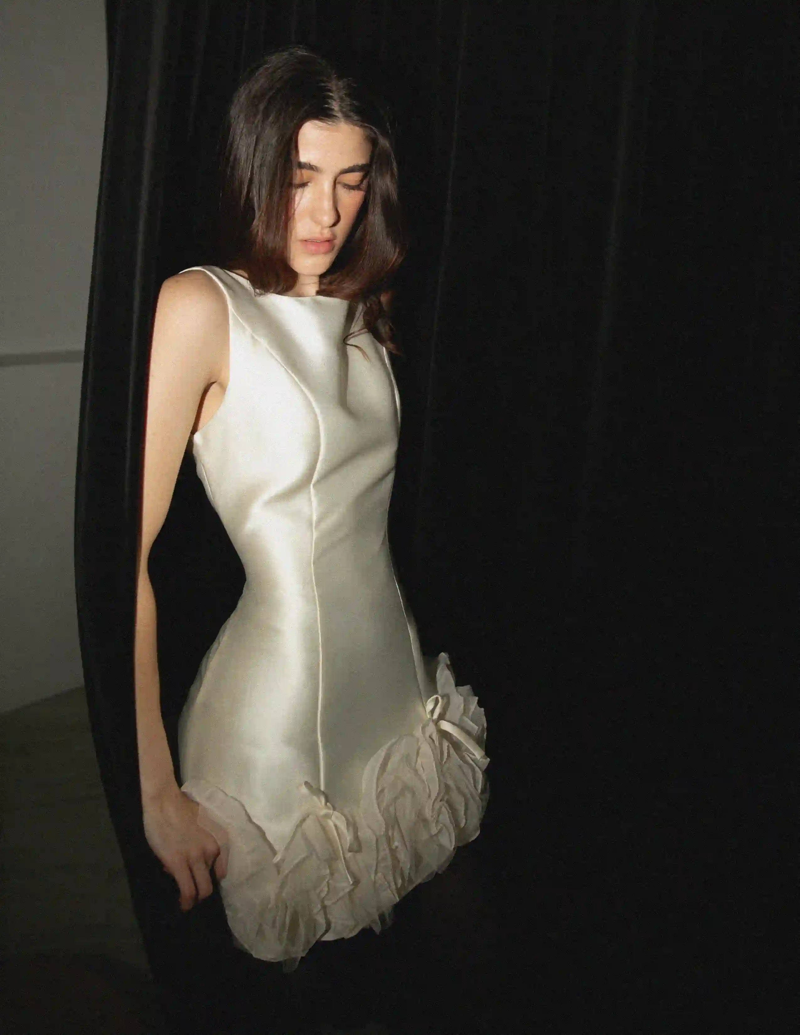 Modelo con vestido blanco satinado de diseño escultural sobre fondo negro, iluminación lateral dramática.