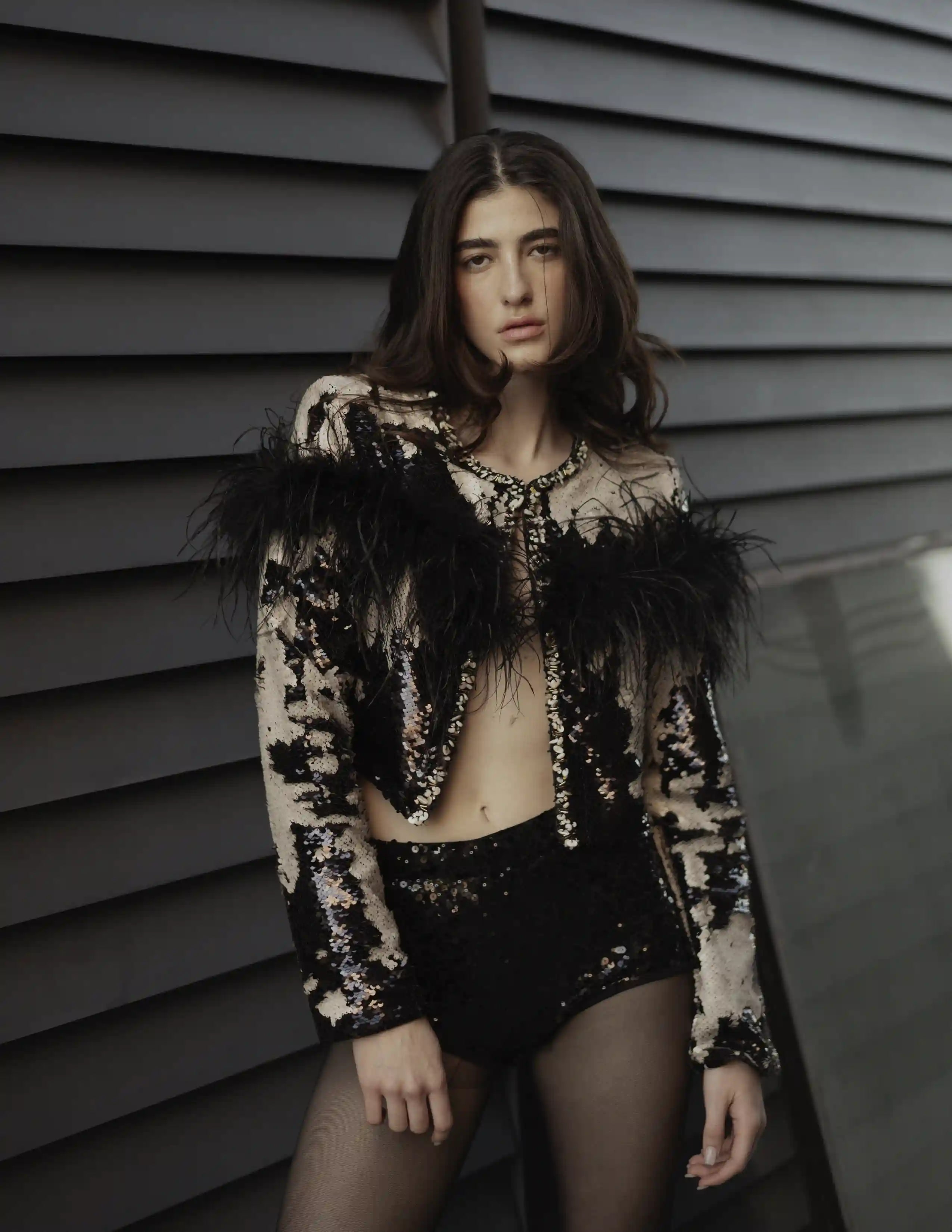 Modelo con chaqueta de lentejuelas bicolor y plumas negras posando sobre fondo industrial gris.