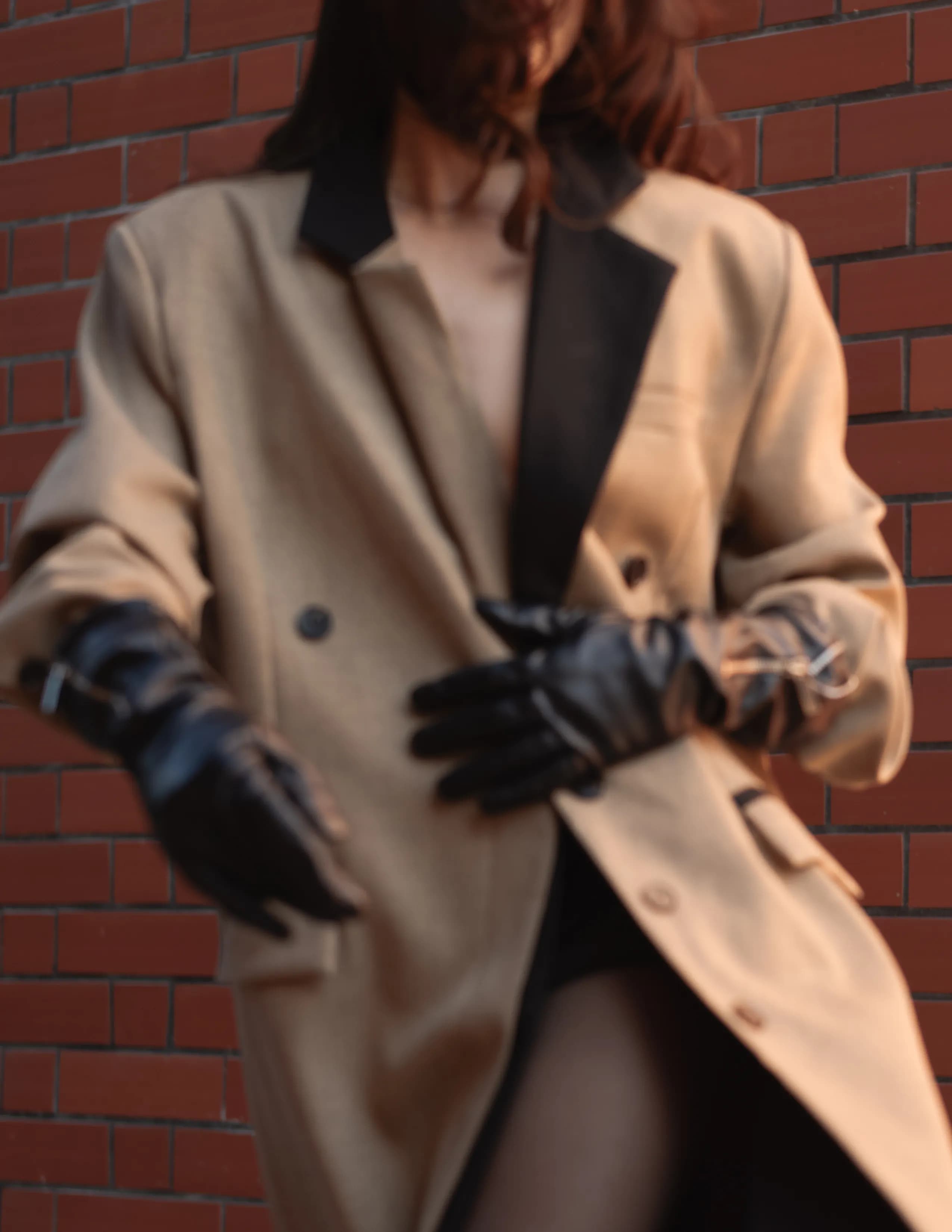 Mujer con trench coat beige y guantes negros sosteniendo gafas de sol contra pared de ladrillos.