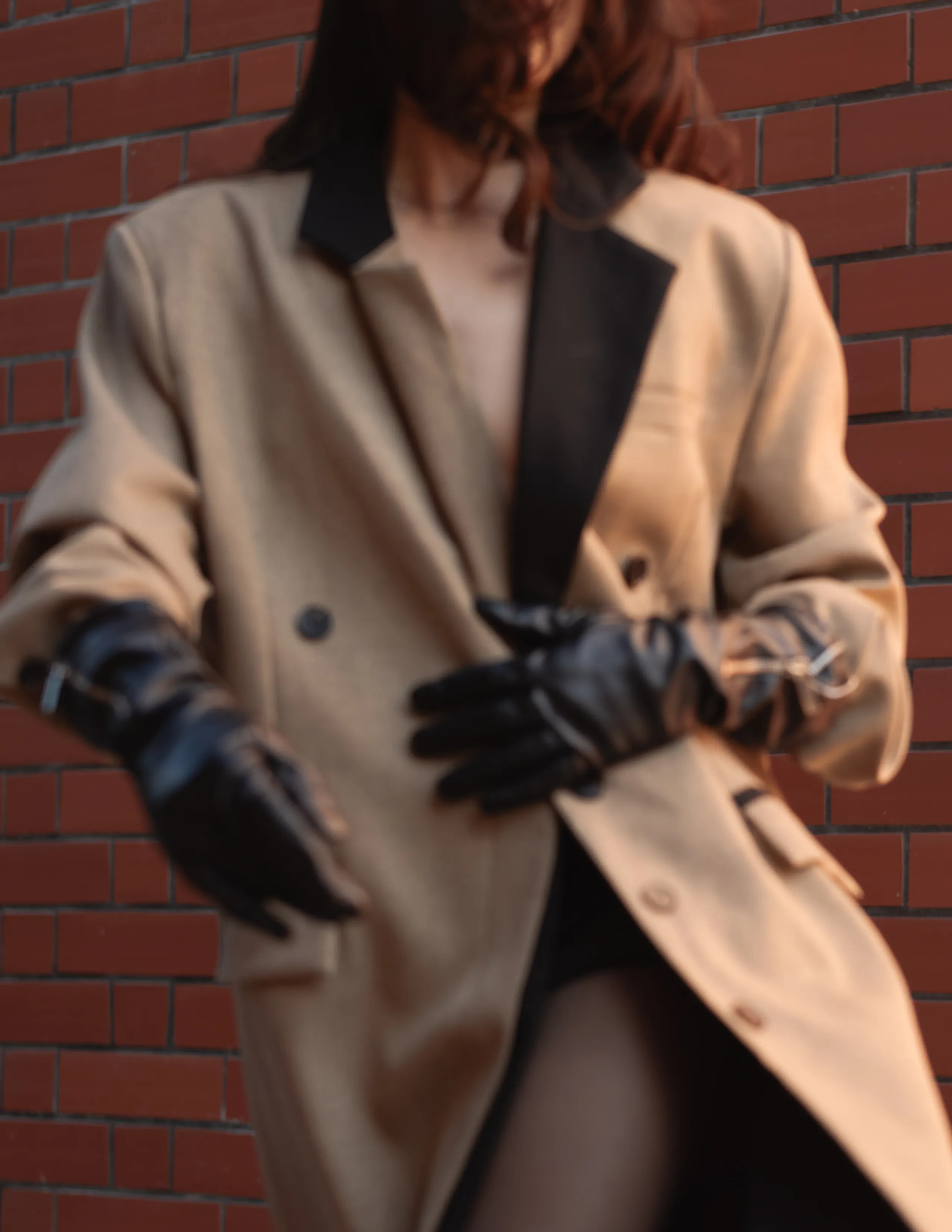 Mujer con trench coat beige y guantes negros sosteniendo gafas de sol contra pared de ladrillos.