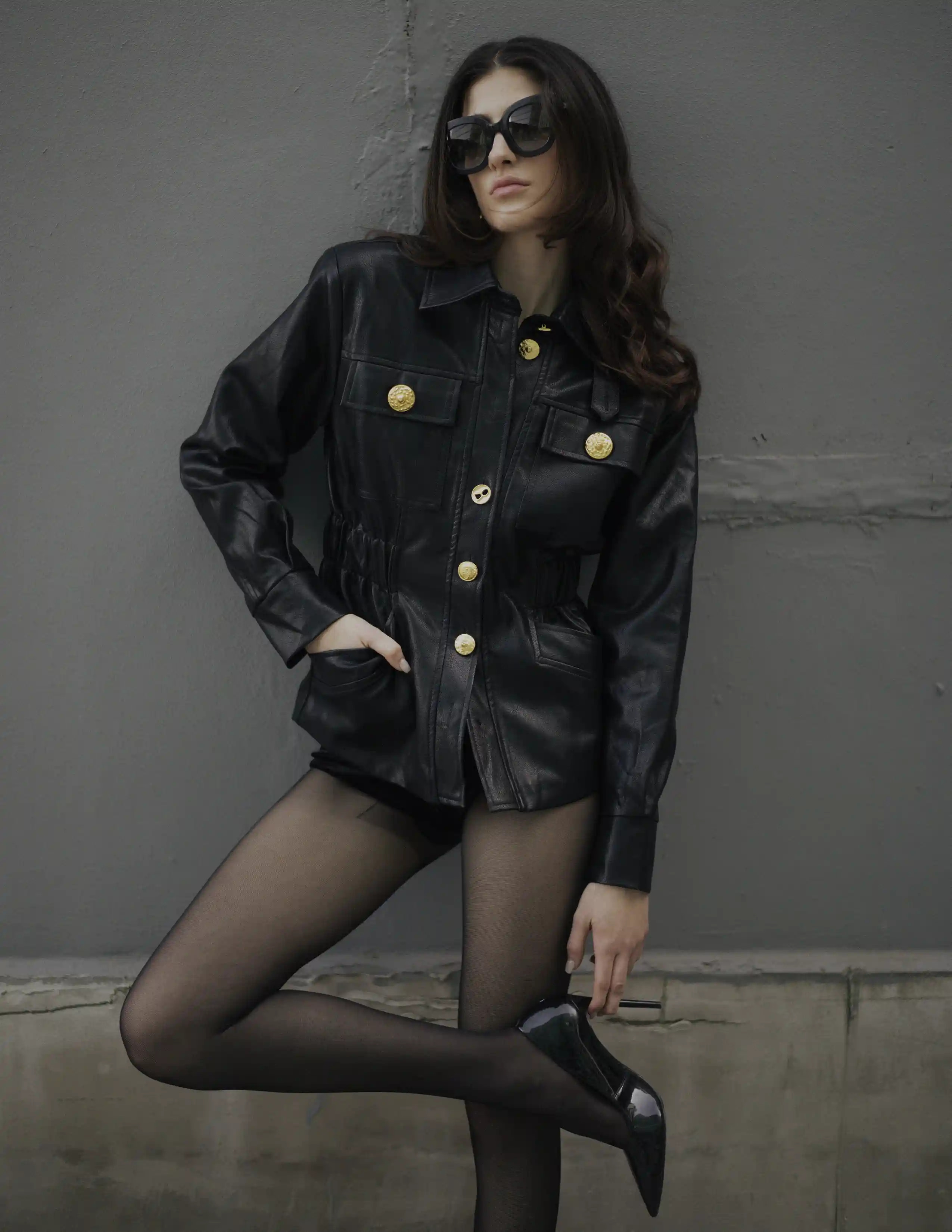 Modelo con chaqueta de cuero negra, medias y tacones posando contra pared gris — moda urbana elegante.