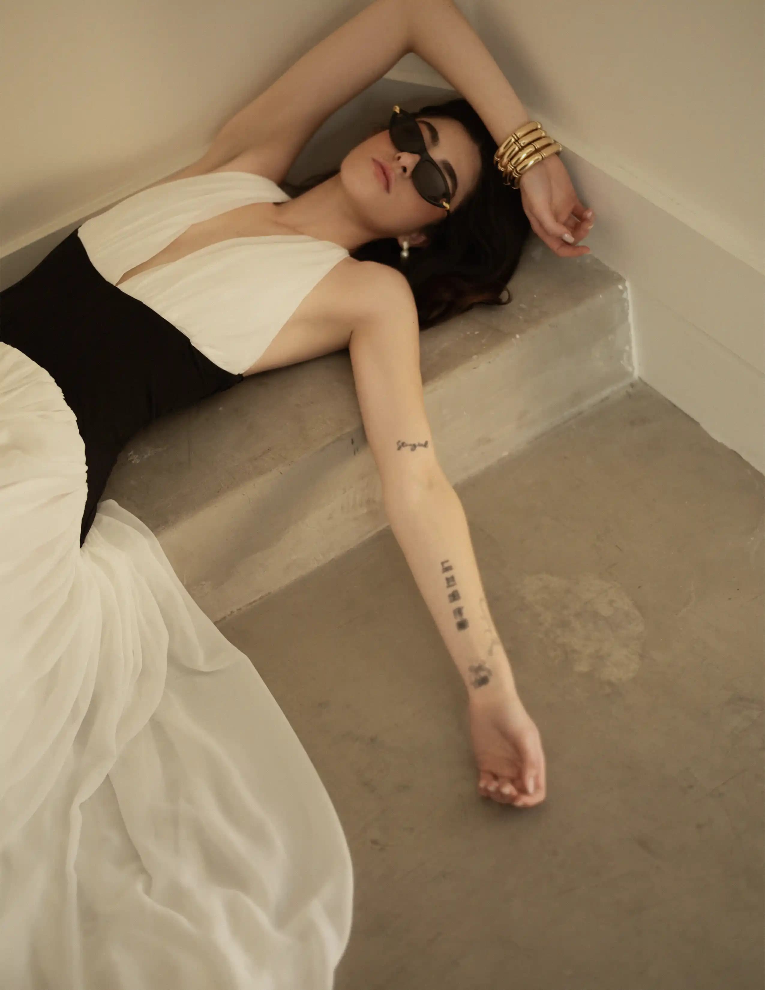 Mujer con vestimenta blanca y negra recostada en escaleras de cemento, gafas de sol y accesorios dorados, estética editorial minimalista.